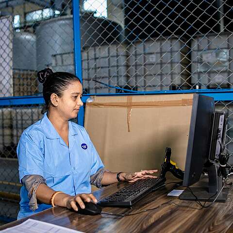 Eine Frau in blauer Uniform sitzt an einem Schreibtisch und arbeitet an einem Computer in einer Industrieanlage. Hinter ihr sind durch einen Metallzaun große Lagerbehälter sichtbar. Auf dem Schreibtisch liegen ein Ventilator und einige Dokumente.