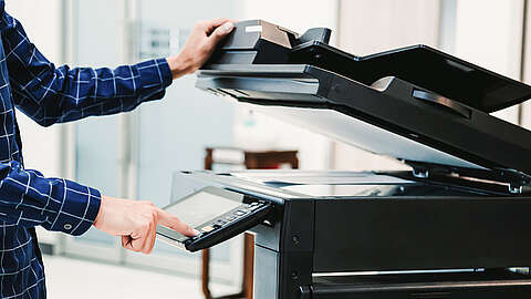Eine Person in einem blau karierten Hemd bedient einen großen schwarzen Drucker, wobei sie mit einer Hand Tasten auf dem Bedienfeld drückt und mit der anderen den Deckel des Druckers anhebt.