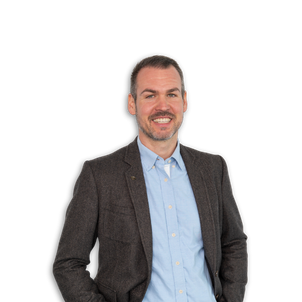 Mann mit kurzen Haaren und Bart, der einen dunklen Blazer über einem hellblauen Hemd trägt, lächelt und vor einem schlichten weißen Hintergrund steht.